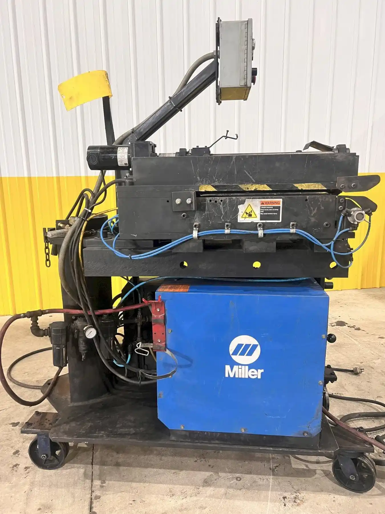 A blue Miller welding power source on a wheeled cart against the yellow and white shop wall